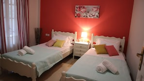 A hotel room with two single beds, each with a white headboard, a bedside table with a lamp, and a floral artwork on the wall.