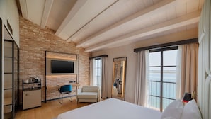 A hotel room with a brick wall, wooden ceiling beams, a bed, a desk with a chair, a TV, and a window with curtains. - NH Collection Venezia Murano Villa (Venice)