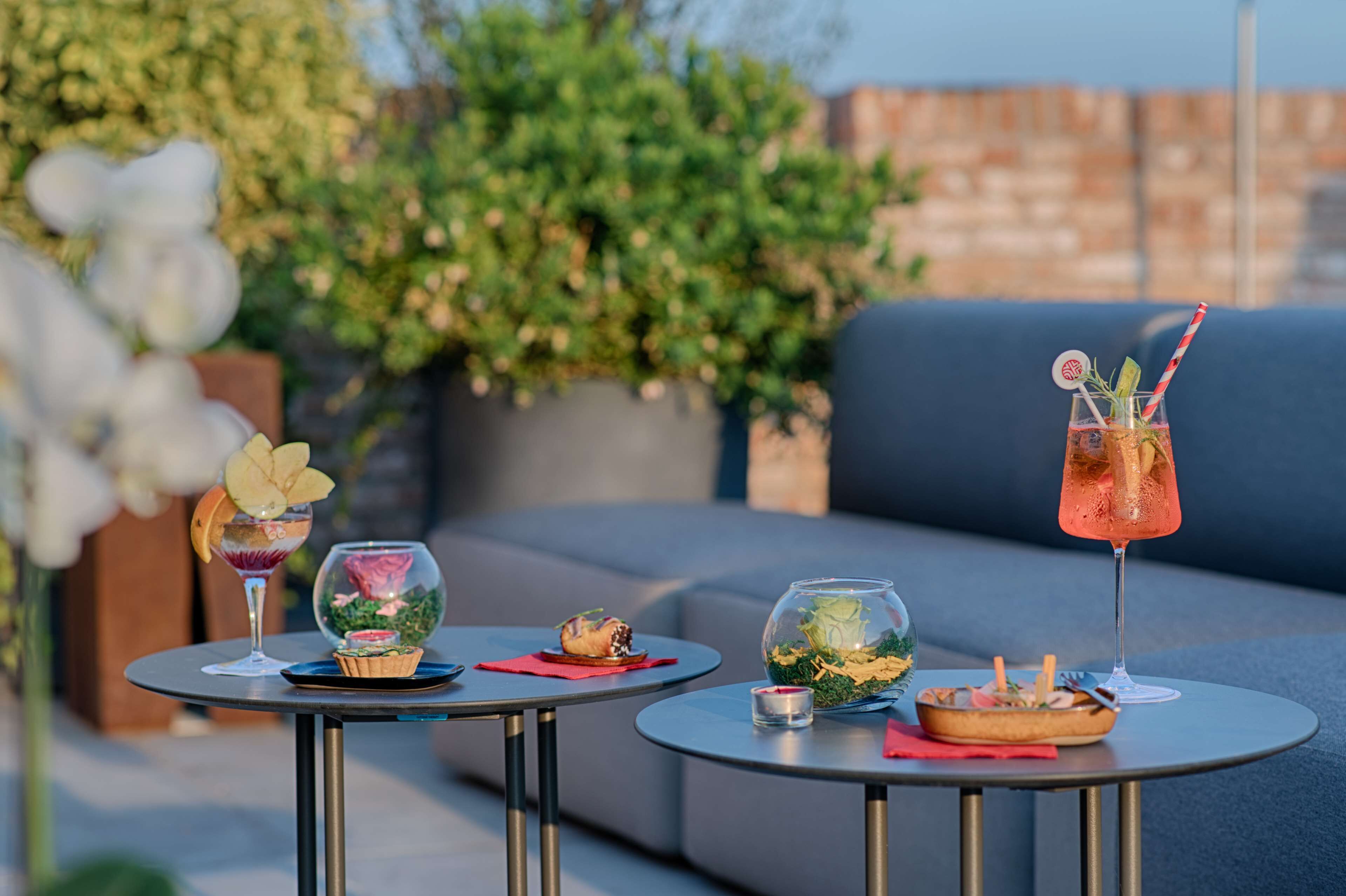 Dos mesas redondas con bebidas y aperitivos, un sofá azul y follaje al fondo.