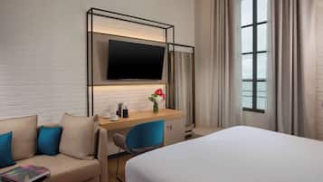 A hotel room with a bed, a sofa with blue cushions, a desk with a flat-screen TV, a coffee maker, and a vase with flowers.
