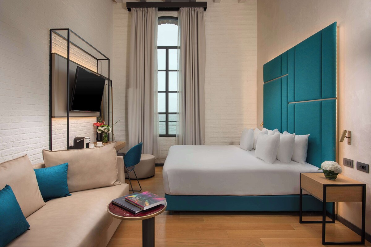 A modern hotel room with a large bed, a sofa, a TV mounted on the wall, and a window with curtains.