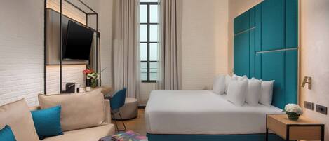 A modern hotel room with a large bed, a sofa, a TV mounted on the wall, and a window with curtains.