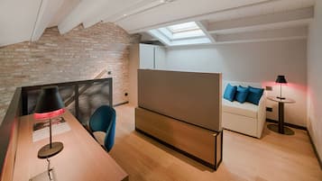 A modern loft bedroom with exposed brick walls, a skylight, a wooden desk, a sofa, and a small round table.