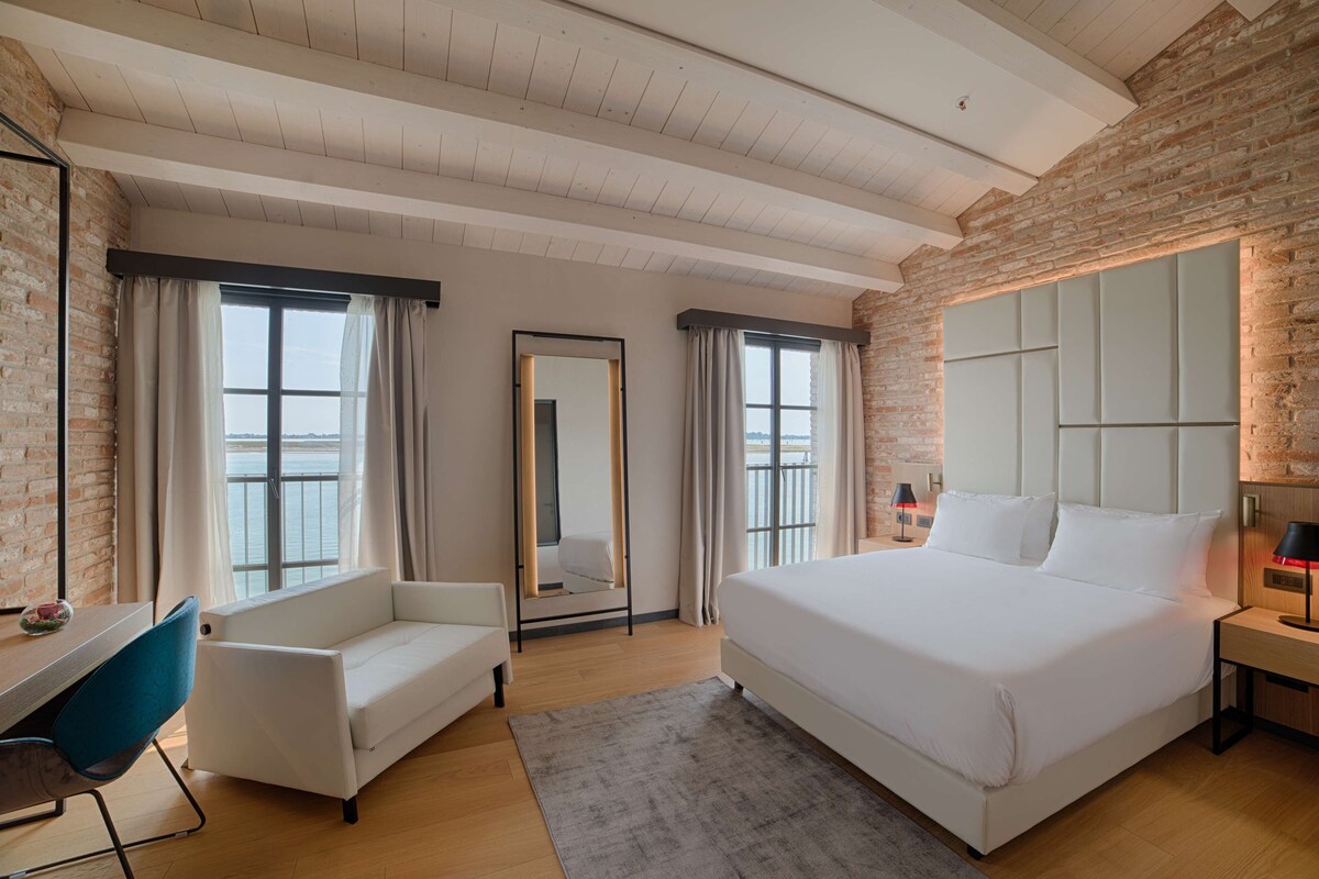 A modern hotel room with a large bed, a desk, a chair, and a view of the sea through large windows.