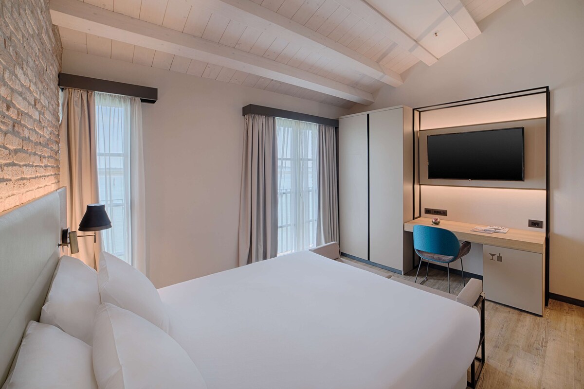 A hotel room with a large bed, a desk with a chair, a flat-screen TV, and a window with curtains.