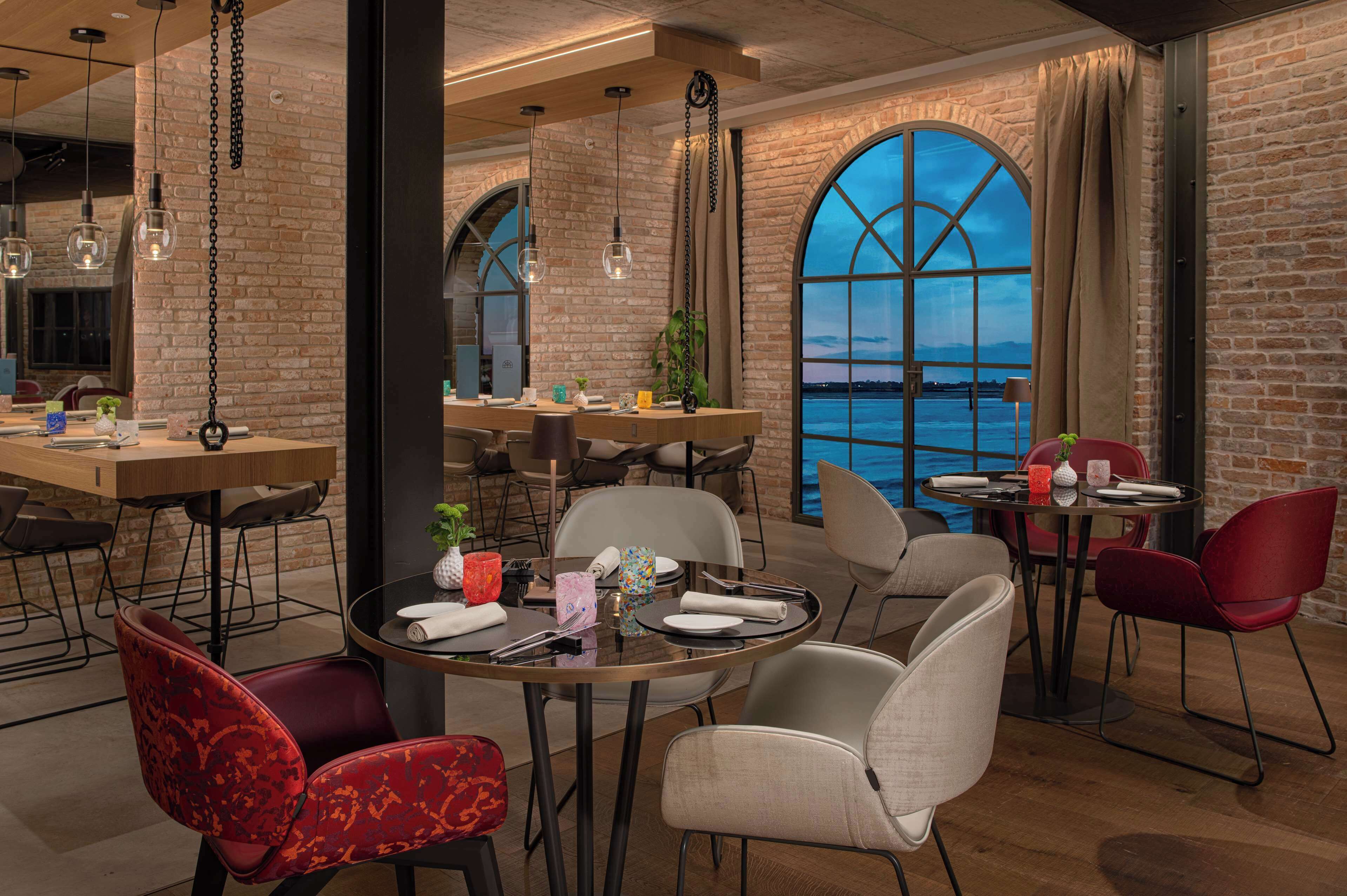Restaurante com parede de tijolos, amplas janelas em arco e luminárias pendentes. Mesas montadas com pratos, talheres e guardanapos.