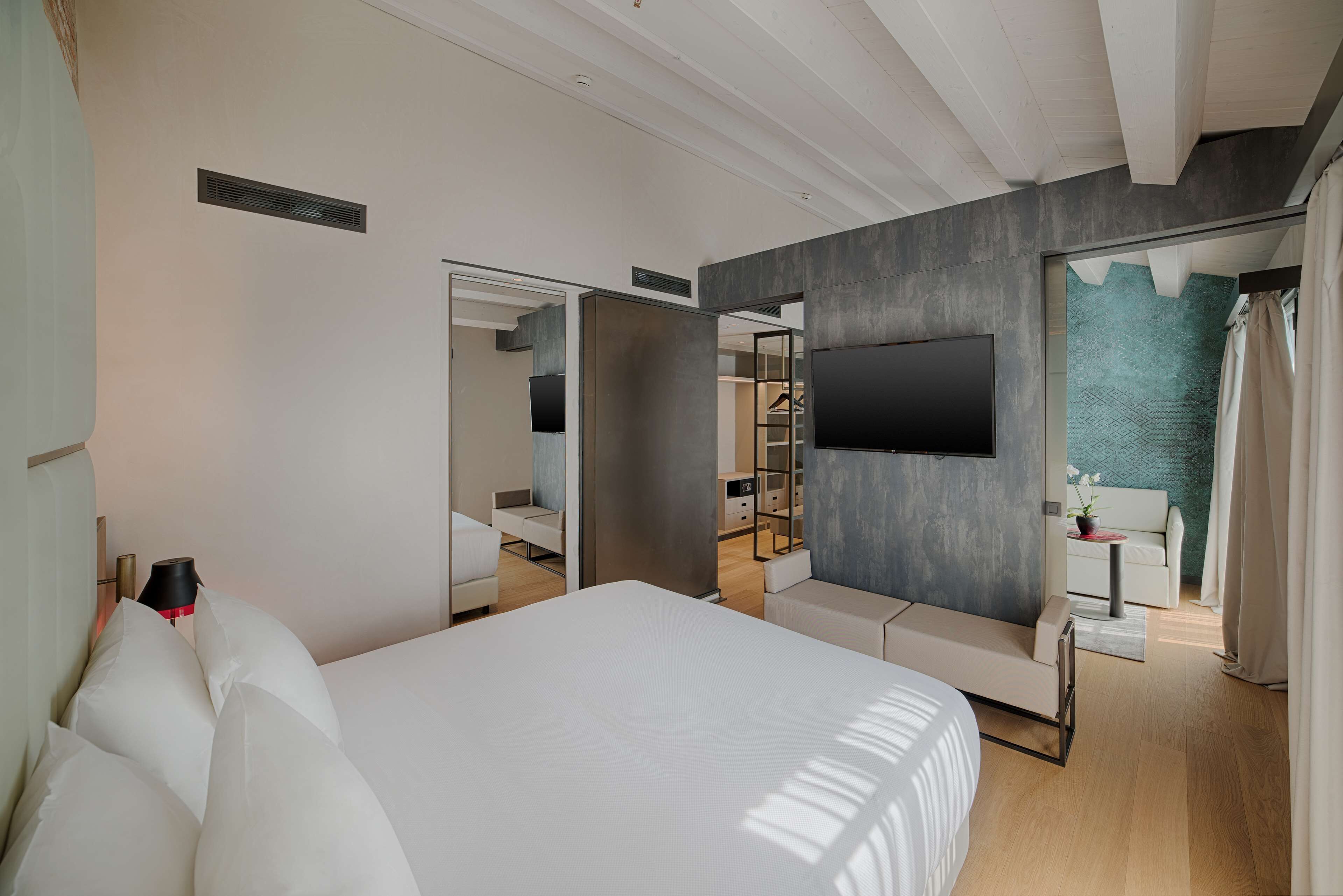 A modern hotel room with a large bed, a TV mounted on the wall, and a view of the sea through a glass door.