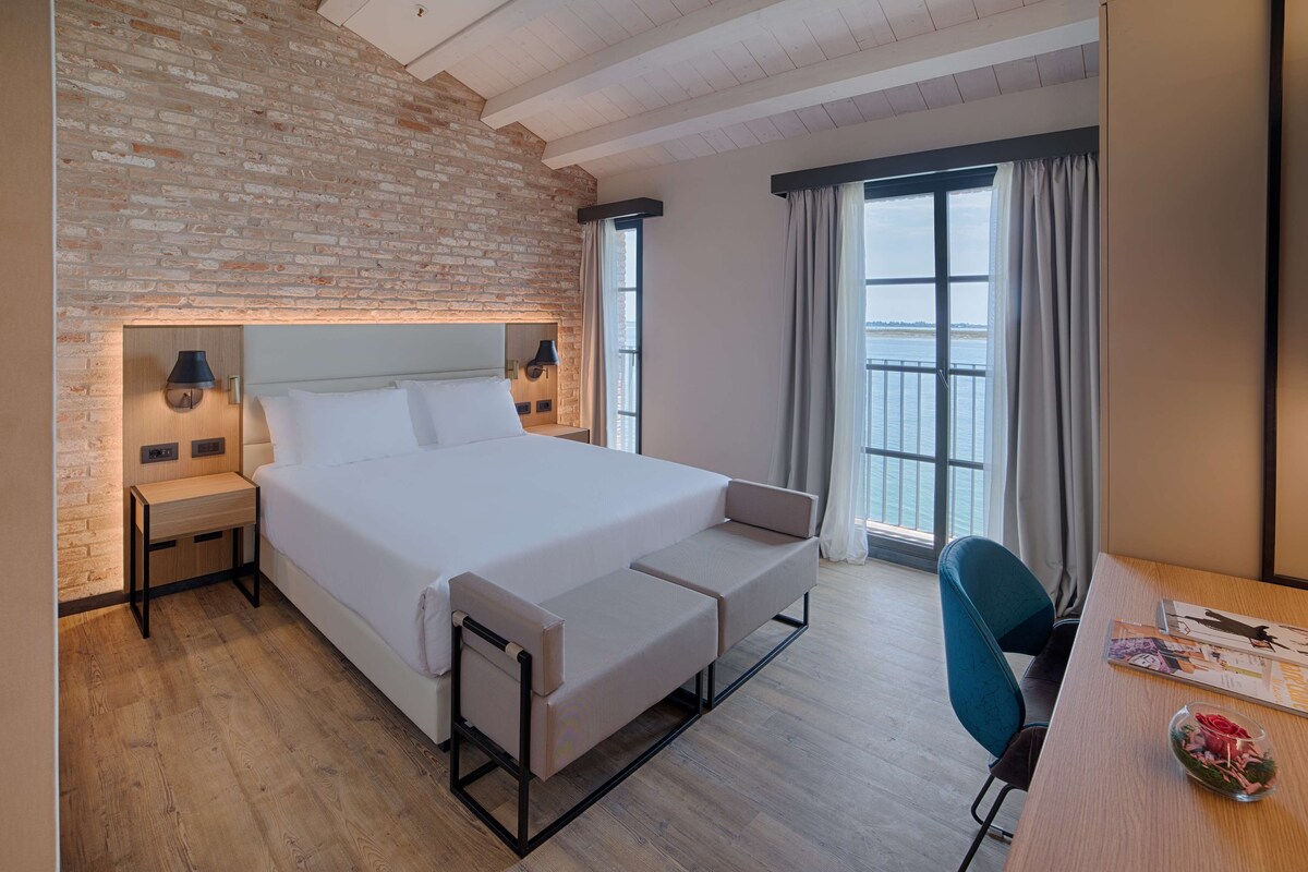 A modern hotel room with a large bed, bedside tables, a desk, and a view of the water.