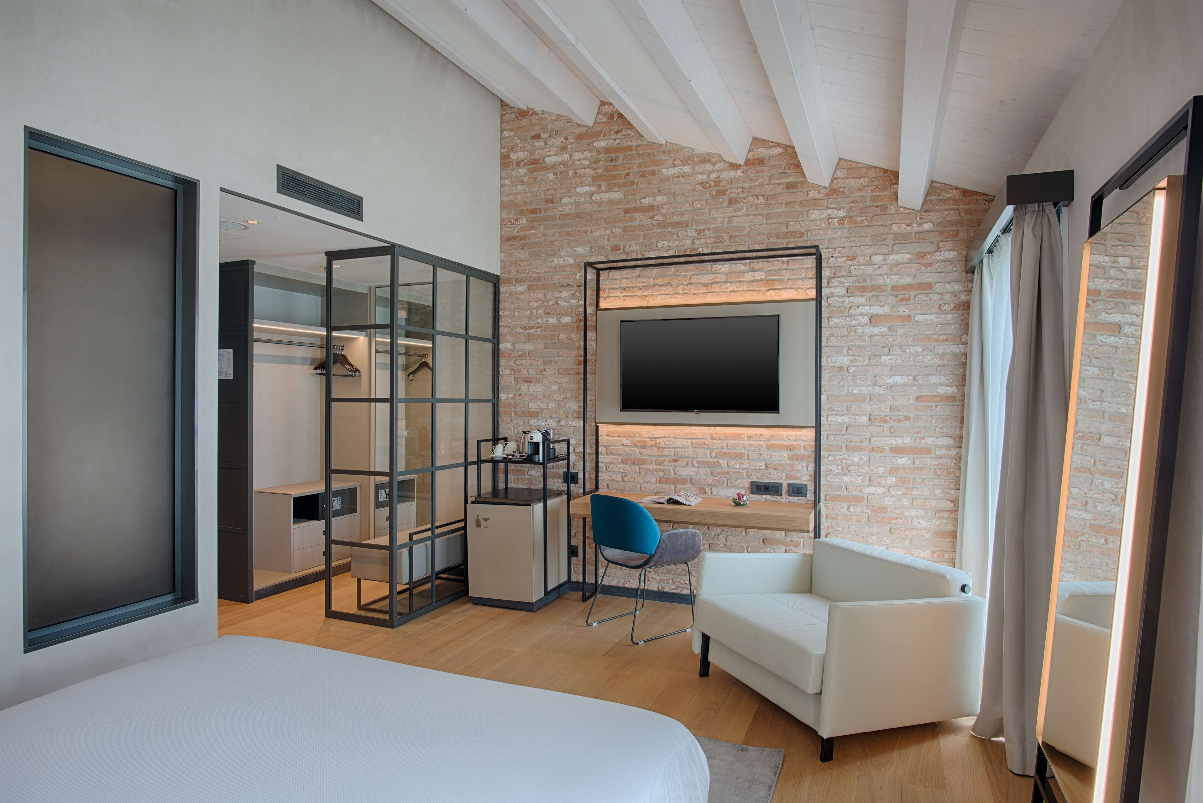 Une chambre d’hôtel moderne avec un mur en briques, un téléviseur à écran plat fixé au-dessus d’un bureau, un canapé blanc et un lit.
