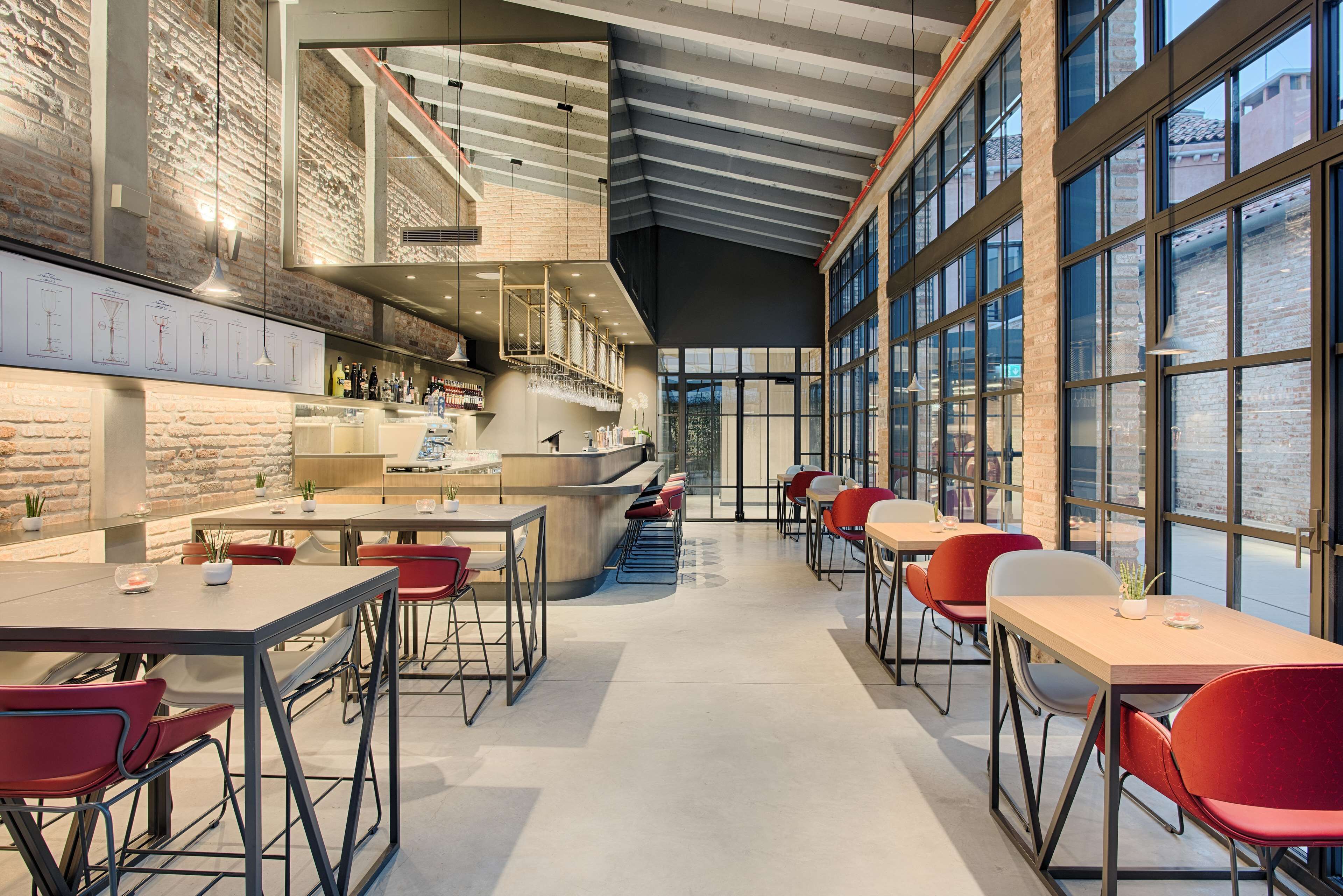 Ein modernes Restaurant mit hohen Decken, freiliegenden Backsteinwänden und großen Fenstern. Tische und Stühle sind zum Essen angeordnet.