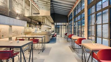 Ein modernes Restaurant mit hohen Decken, freiliegenden Backsteinwänden und großen Fenstern. Tische und Stühle sind zum Essen angeordnet.