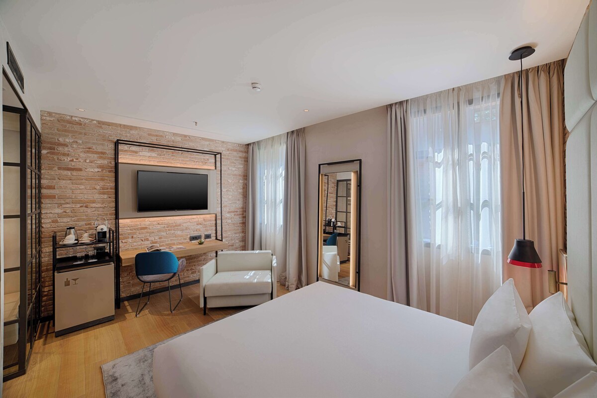 A hotel room with a large bed, a desk with a chair, a TV mounted on the wall, a small kitchenette, and a large window with curtains.