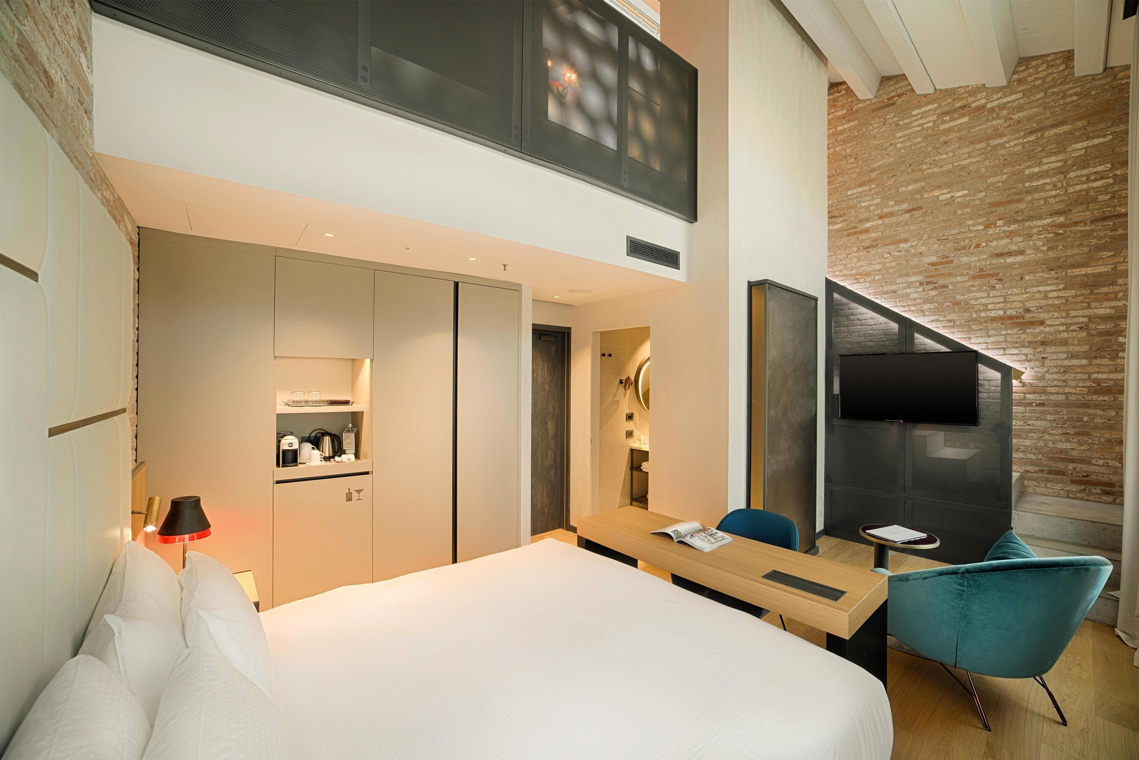 A modern hotel room with a large bed, a desk, a chair, a TV, and a kitchenette.