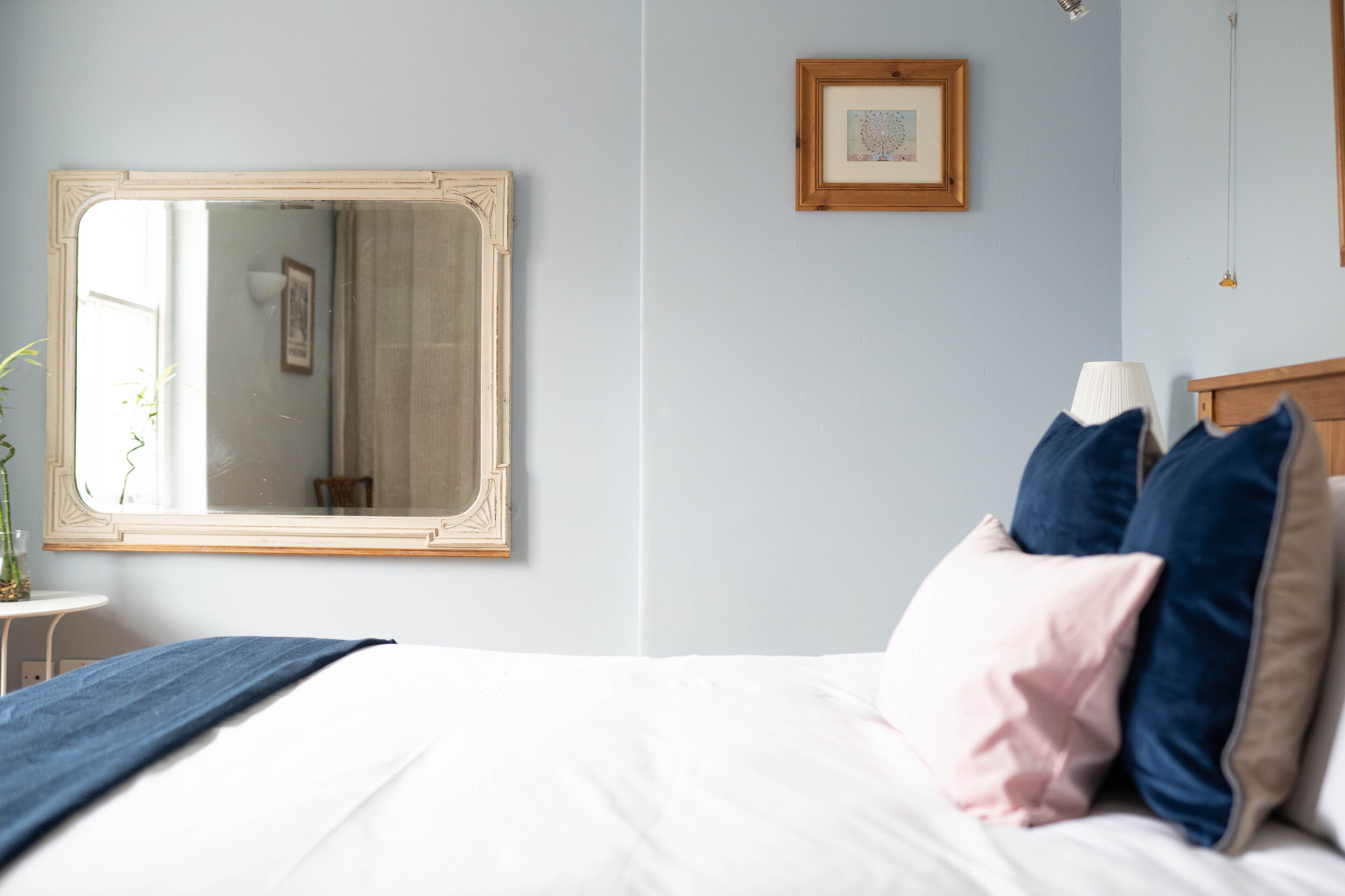 A bedroom with a large mirror, a bed with white bedding, and a small table with a vase.