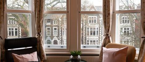 Una habitación con dos sillas, una pequeña mesa con una planta y ventanas grandes con cortinas.