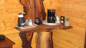 Une table en bois rustique, sur laquelle se trouvent une machine à café, un plateau avec une théière, des tasses et une petite plante. Un fusil est accroché au mur, au-dessus de la table.