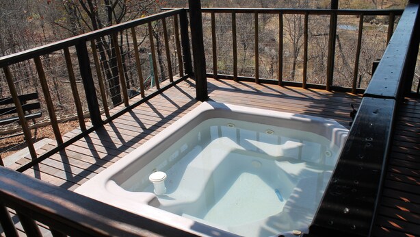 Un bain à remous sur une terrasse en bois, avec une rambarde en métal, entouré d’arbres.