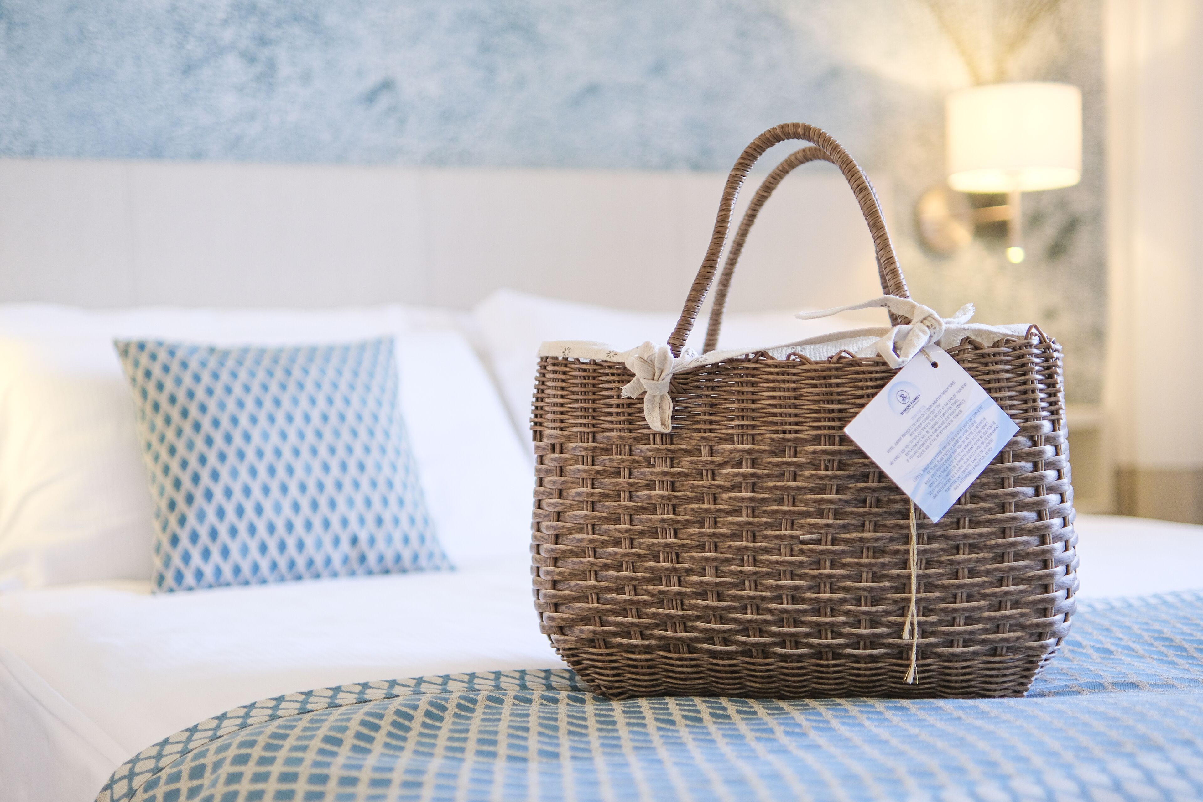 A wicker basket with a tag on a bed with patterned pillows.