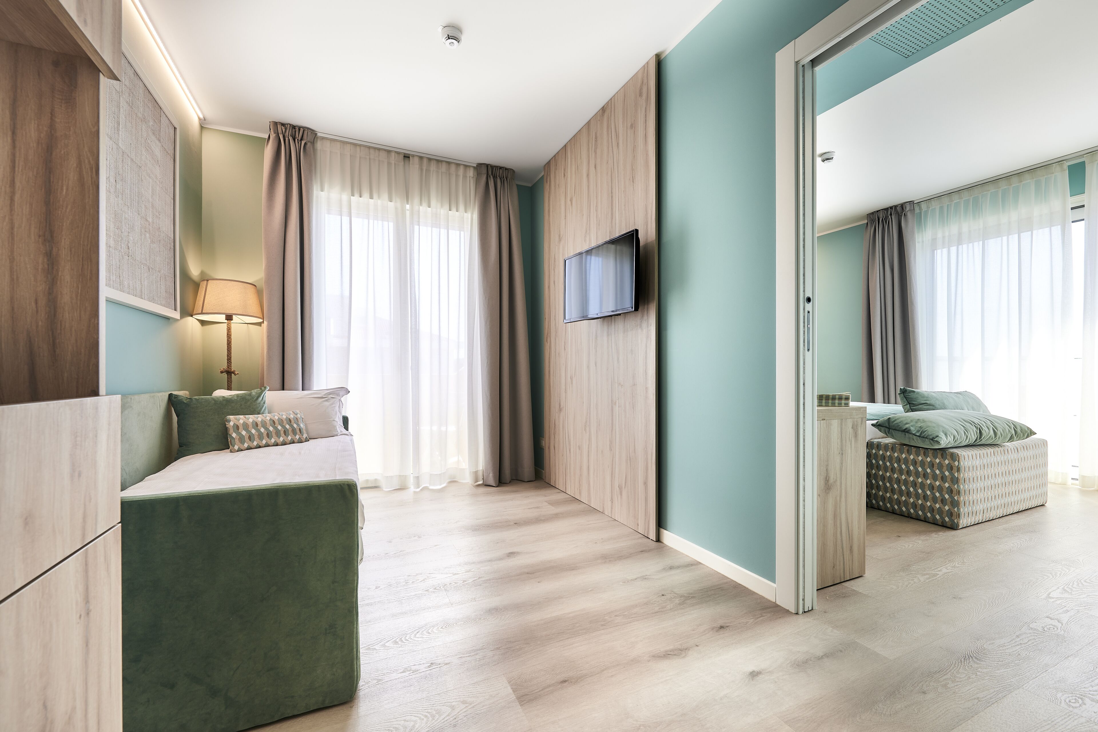 A modern hotel room with a bed, a TV mounted on the wall, and a seating area with a sofa and a table.