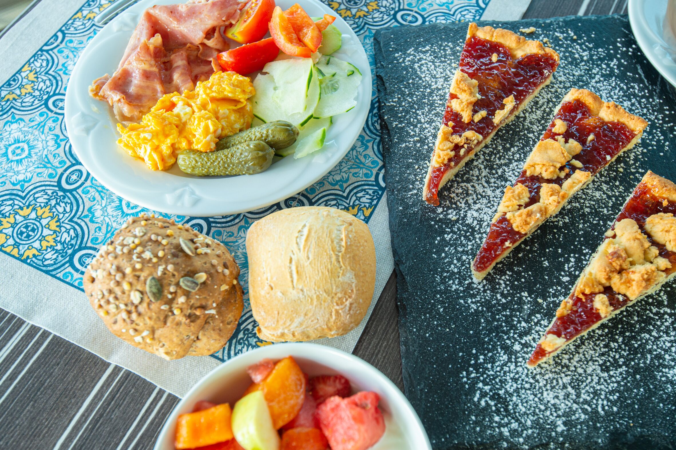 Un plato de desayuno con huevos revueltos, jamón, verduras y una rebanada de tarta.