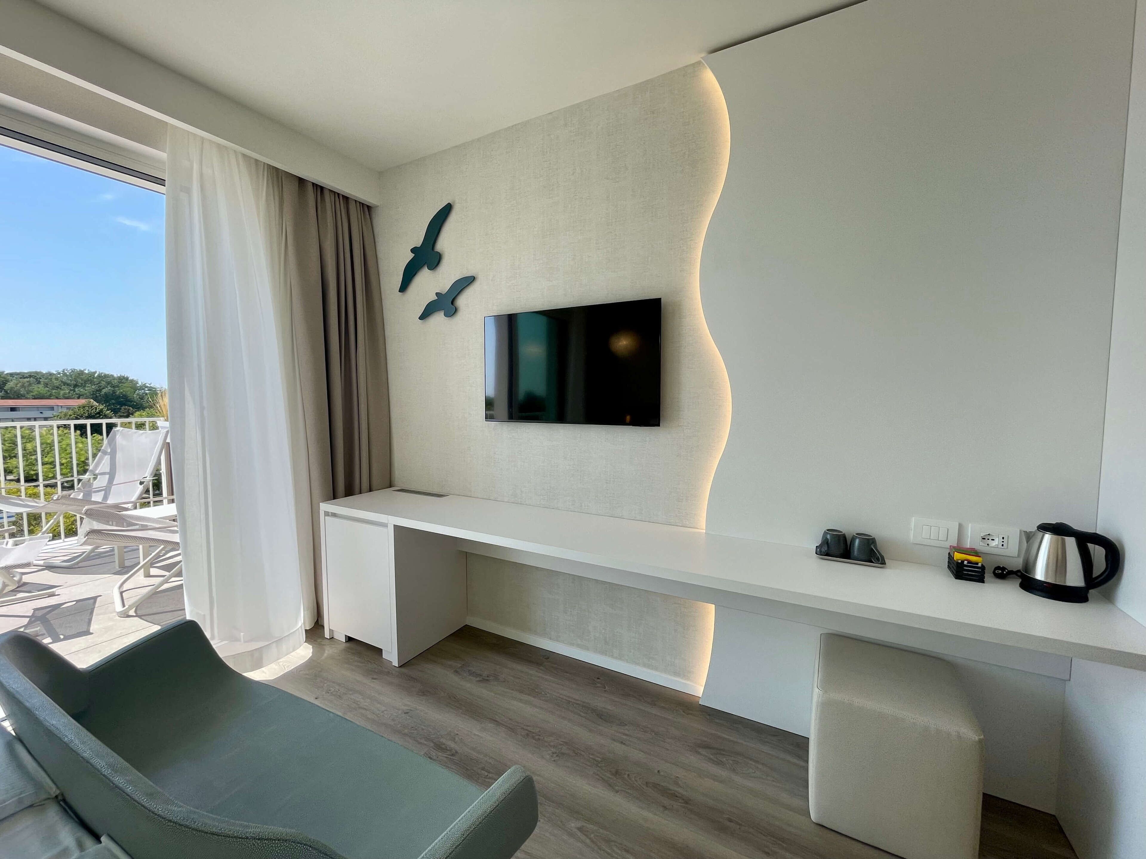 Ein modernes Hotelzimmer mit einem Flachbildfernseher an einer strukturierten Wand, einem weißen Schreibtisch, einem gepolsterten Sessel und Blick ins Freie.