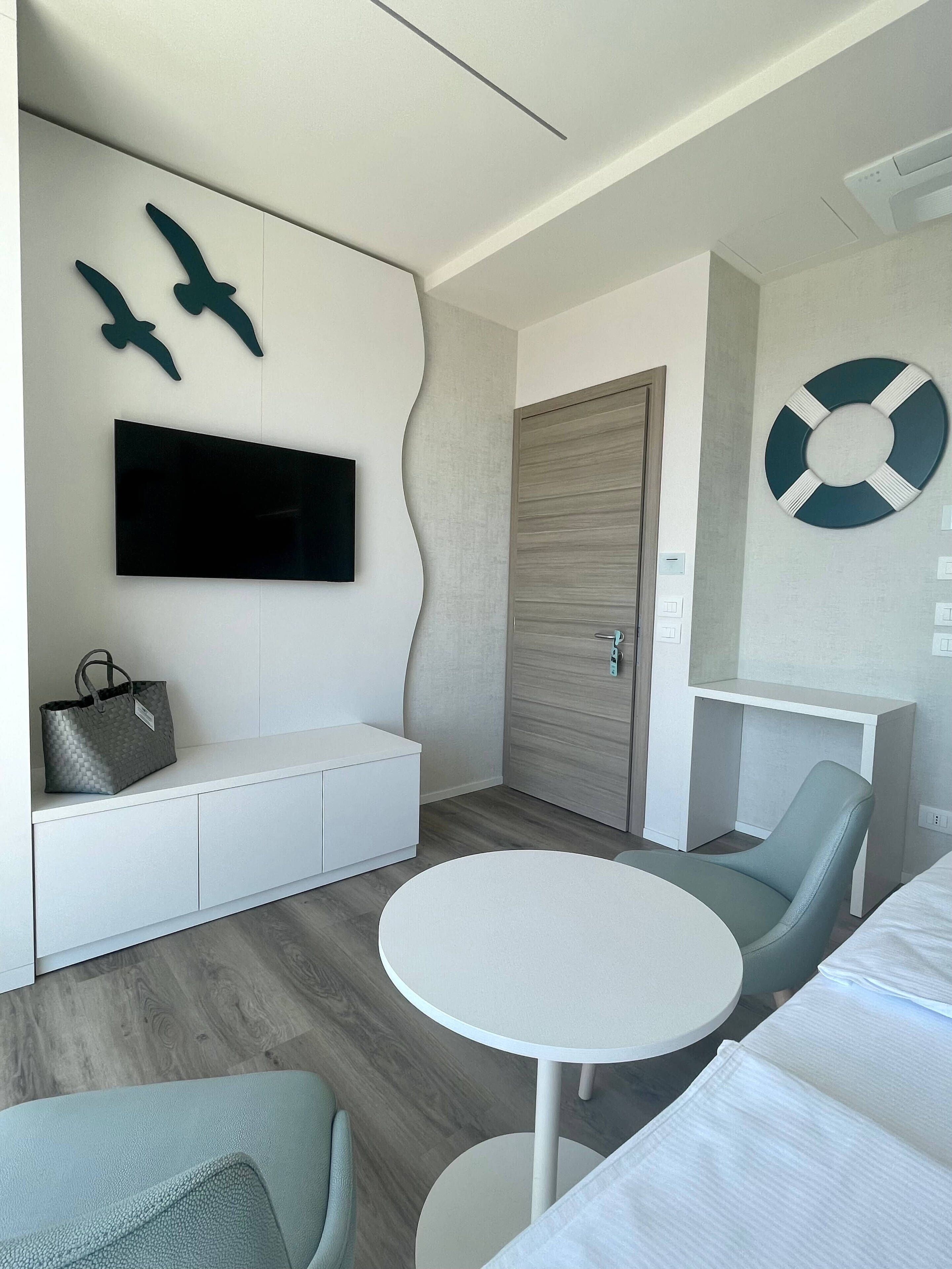 Une chambre d’hôtel moderne équipée d’un téléviseur à écran plat, d’une table ronde, d’un canapé, d’un banc et d’un élément décoratif en forme de bouée fixé au mur.