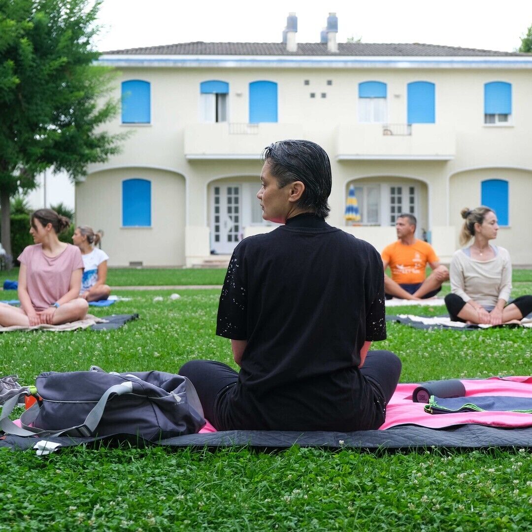 A group of people sitting on mats outdoors in front of a building with blue windows and a white facade.