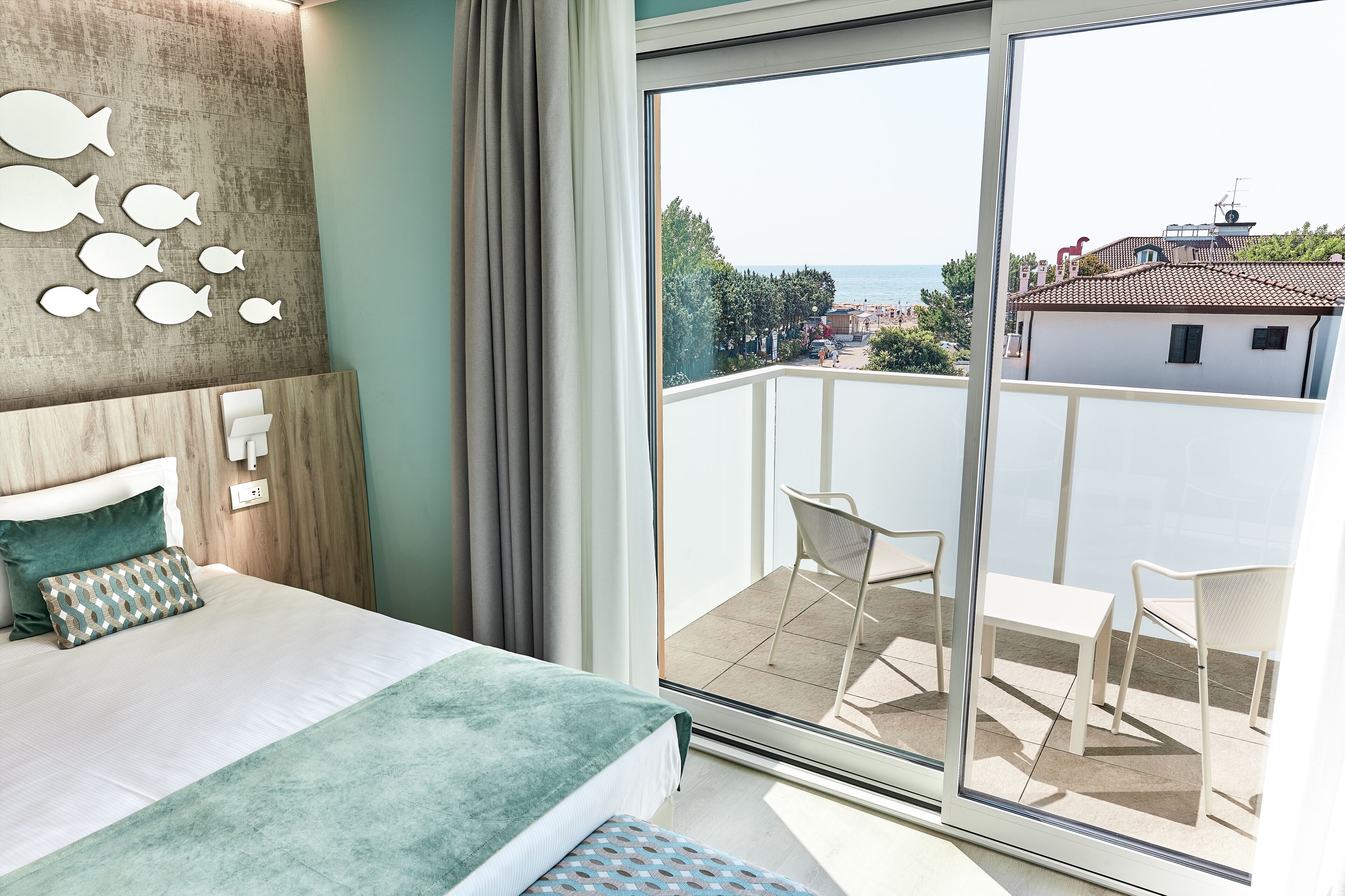 A bedroom with a large bed, a view of the sea through a sliding glass door, and a balcony with chairs.