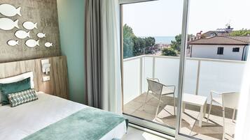 A bedroom with a large bed, a view of the sea through a sliding glass door, and a balcony with chairs.