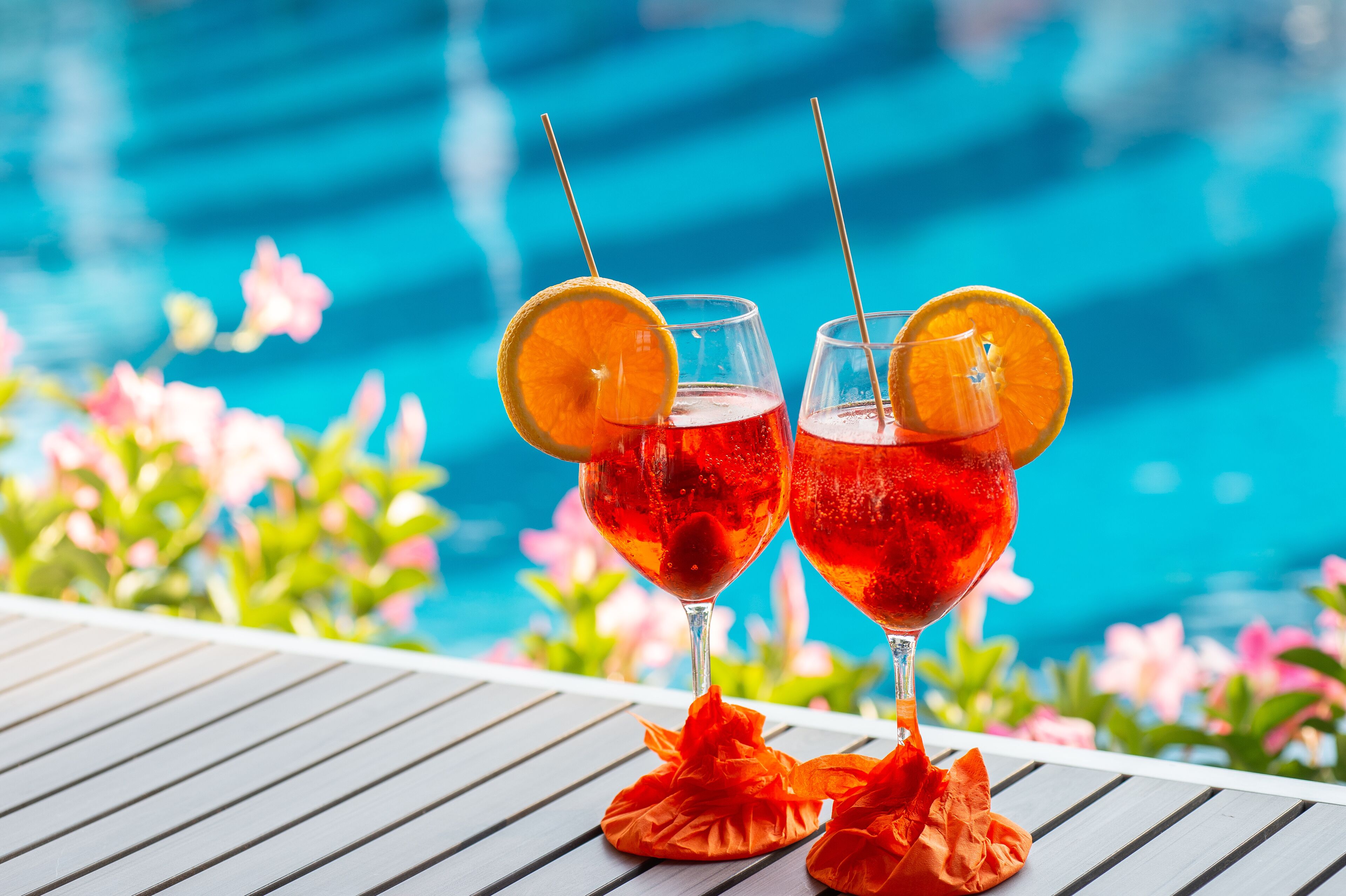 Two glasses of red beverage with orange slices on a table by a poolside with pink flowers in the background.