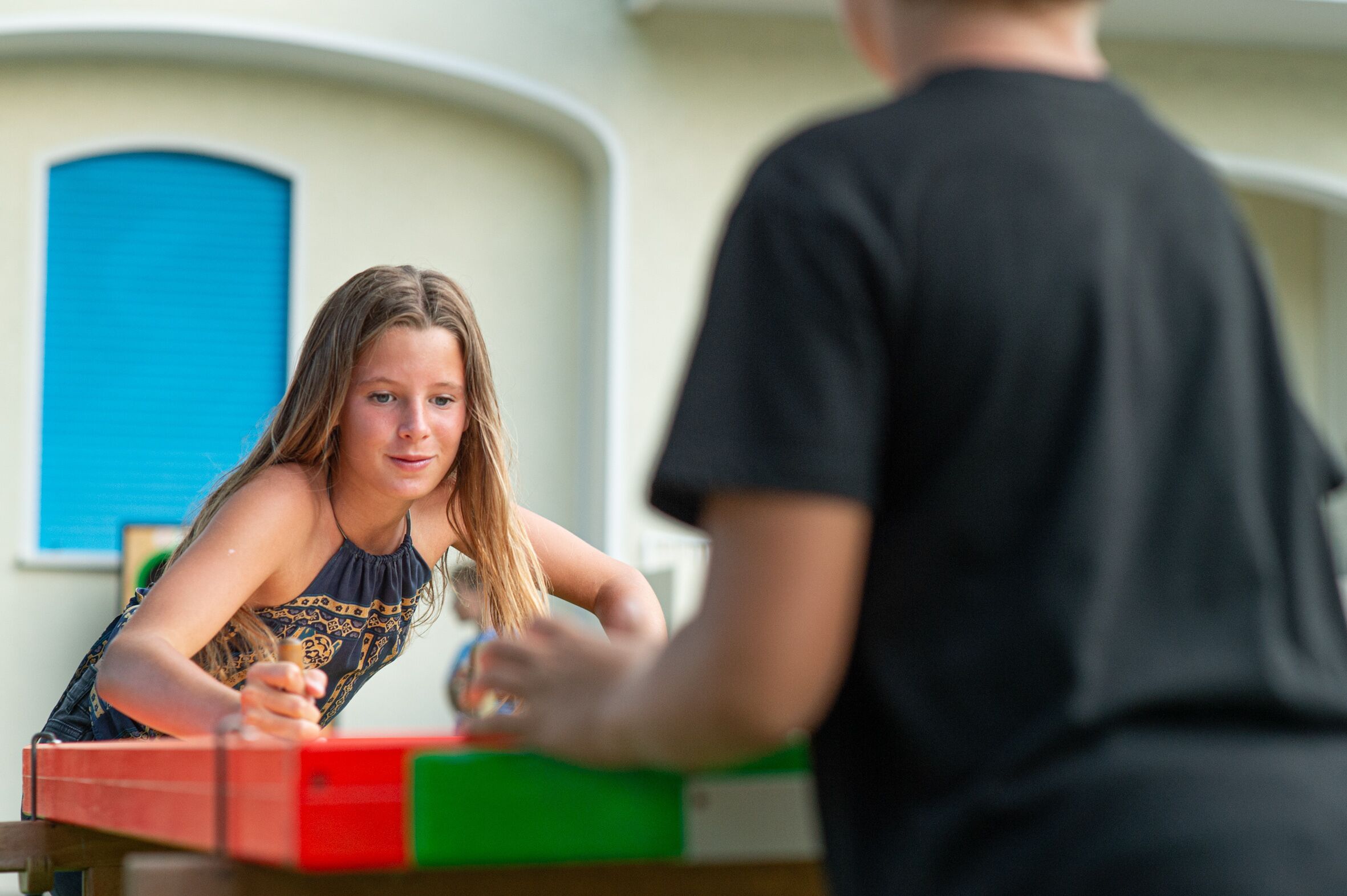Une personne est assise à une table devant un objet rouge et vert, qui pourrait être un jeu de société, dans une pièce avec une porte bleue et un mur blanc.