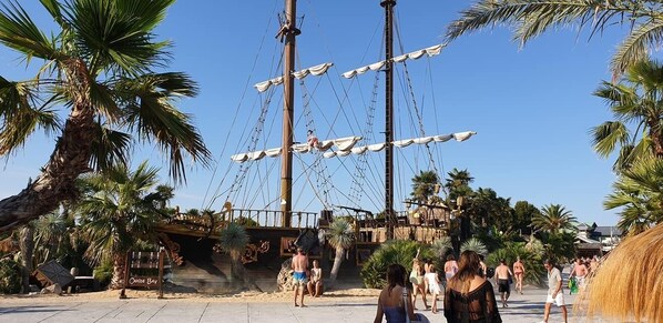 Un paseo marítimo en la playa con un mástil de barco grande, palmeras y gente caminando.