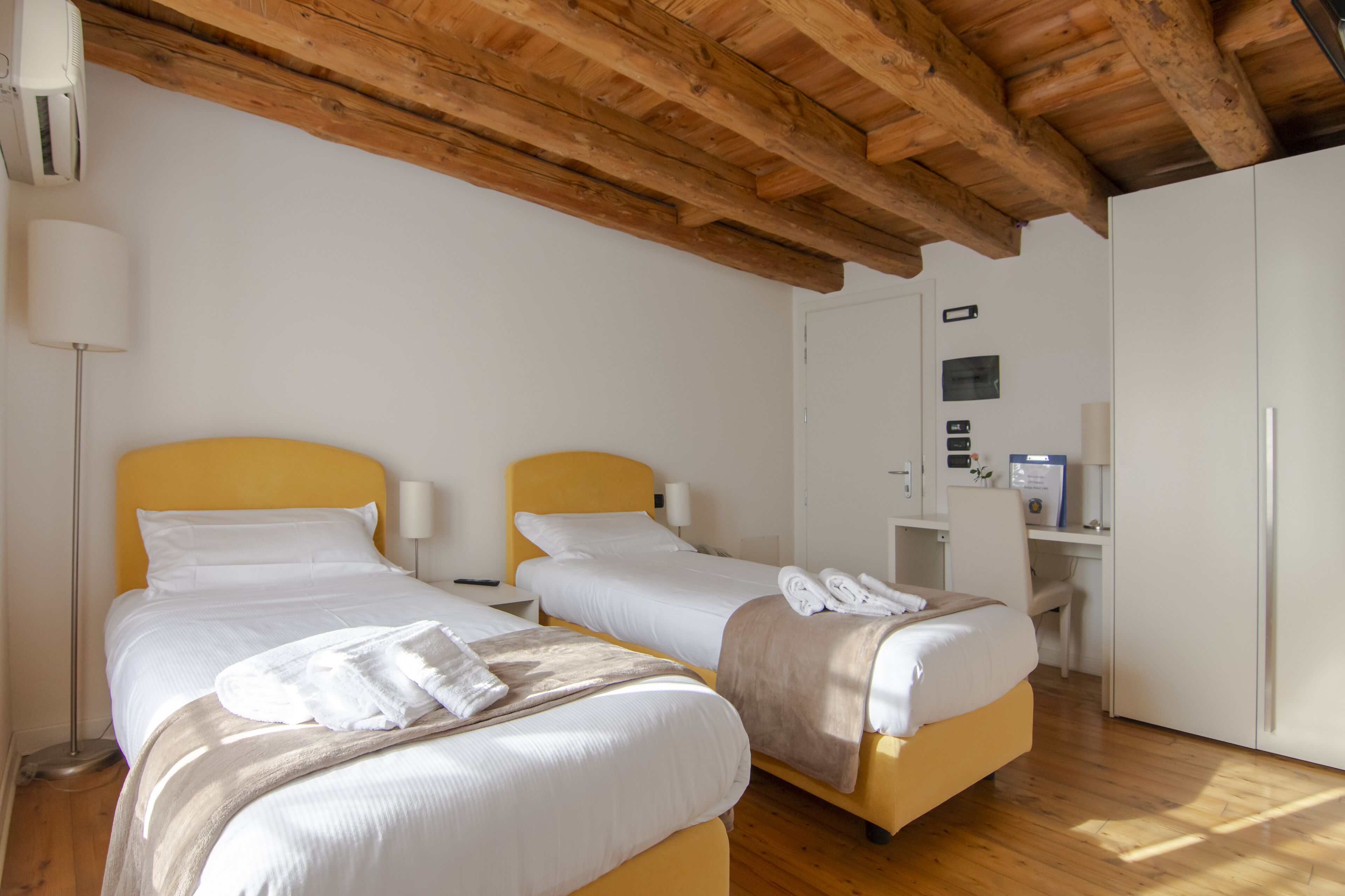 A hotel room with two beds, wooden ceiling beams, a desk with a computer, and a wardrobe.