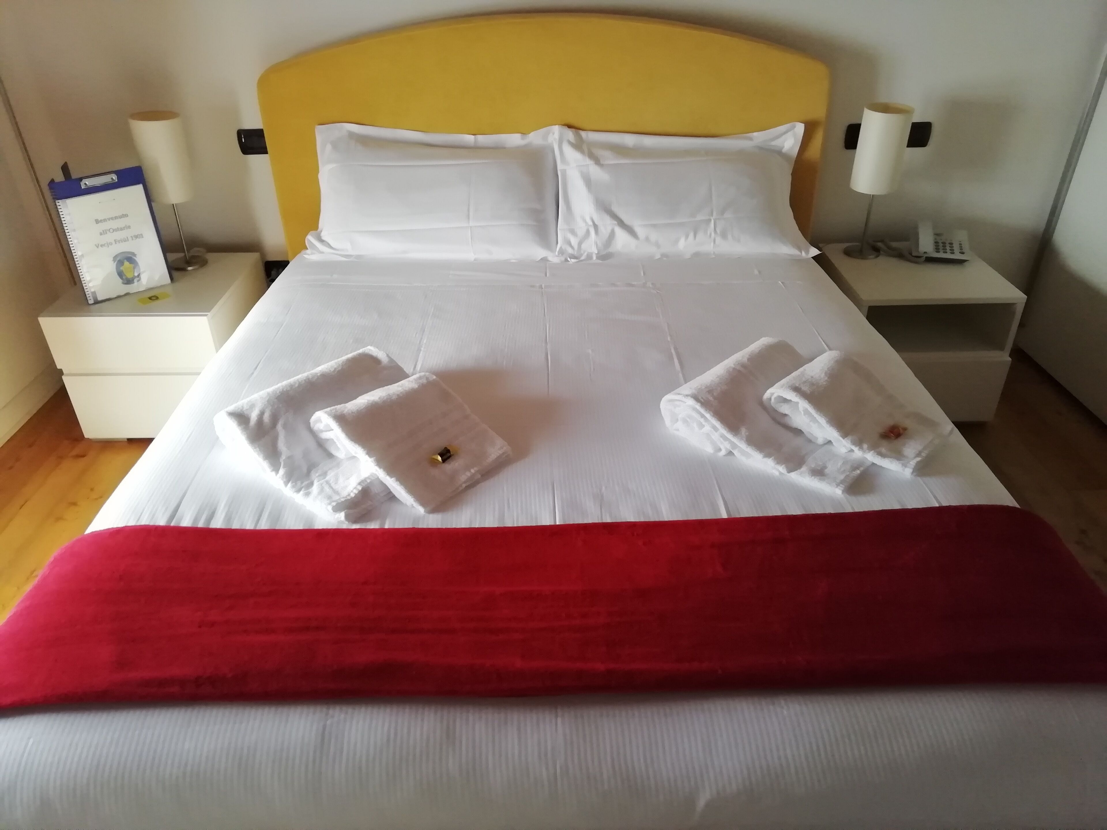 A neatly made bed with white linens, two folded towels, a red blanket, and a yellow headboard.