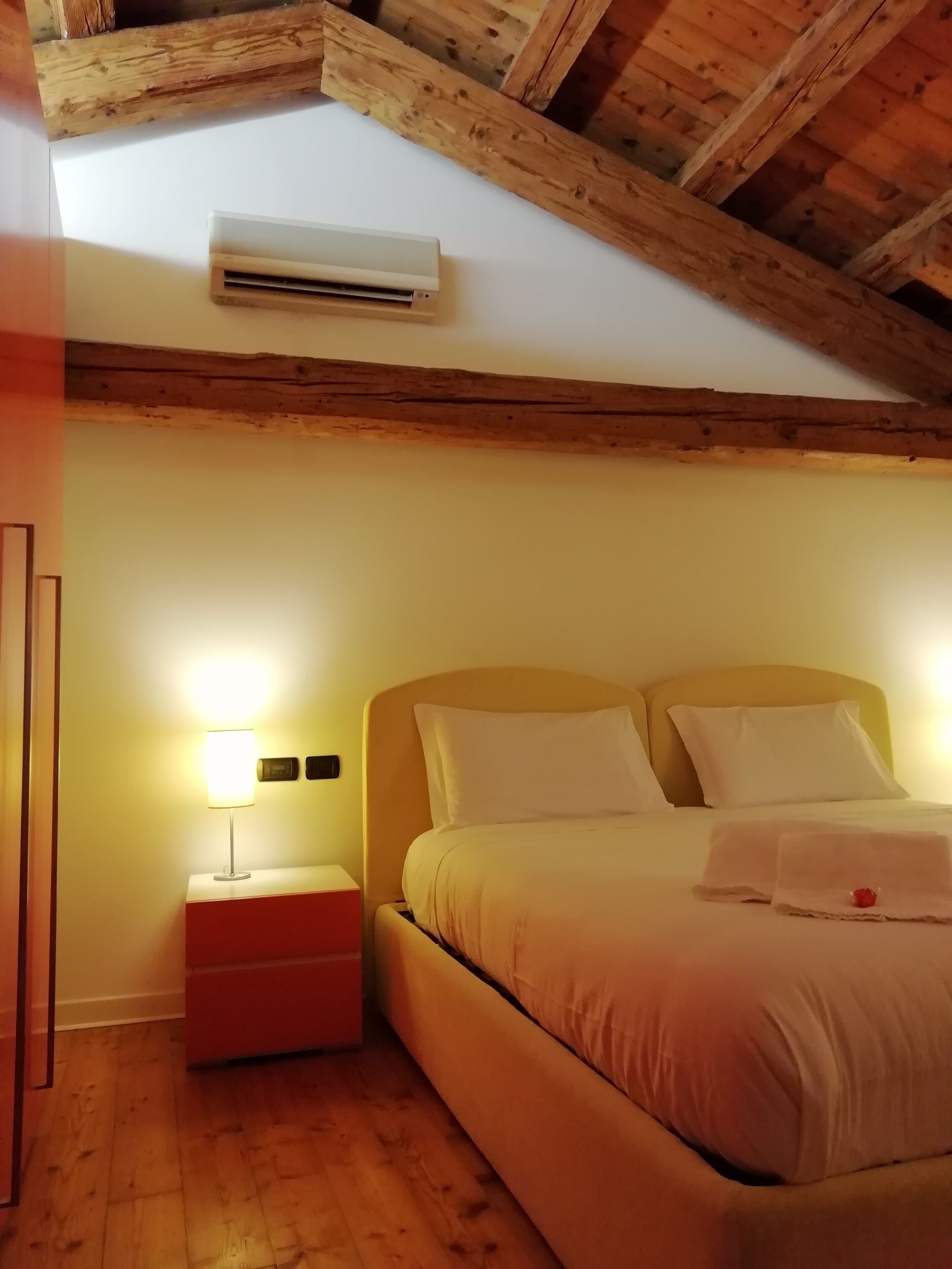 A bedroom with a wooden ceiling, a bed with white bedding, a bedside table with a lamp, and an air conditioning unit.