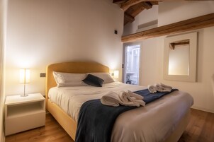 Una cama bien tendida con sábanas blancas y una manta azul oscuro, una lámpara de noche y una toalla doblada sobre la cama.