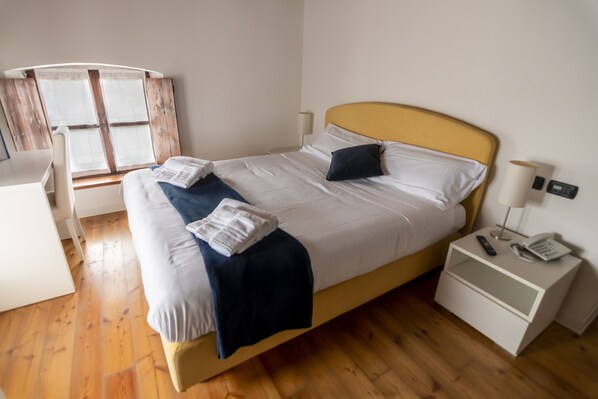 A neatly made bed with white linens, a dark blue throw blanket, and a single dark pillow. There is a bedside table with a lamp and a phone. The room has wooden flooring and a window with blinds. - Ostarie Vecjo Friûl (Sedegliano)