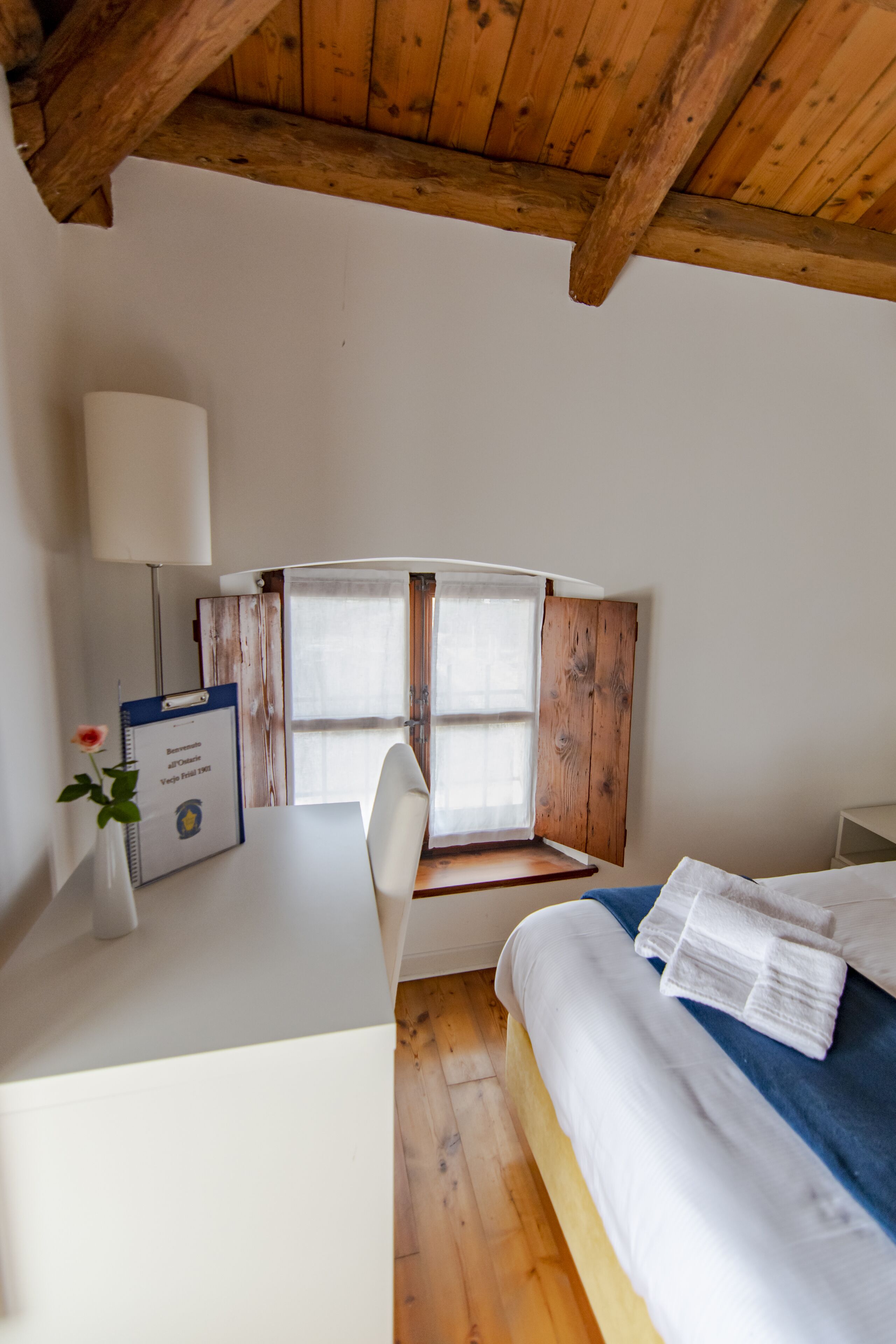 Un dormitorio con techo de madera, cama con ropa de cama blanca, escritorio con lámpara y ventana con persianas de madera.
