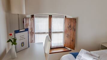 A bedroom with a wooden ceiling, a bed with white bedding, a desk with a lamp, and a window with wooden shutters.