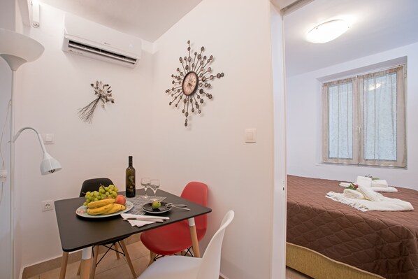 A compact room with a dining table set for two, a bed with a brown quilt, and a decorative wall piece. - House Loukia  by CorfuEscapes (Corfu)