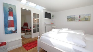 Une chambre à coucher avec un lit, un téléviseur posé sur un meuble, un tableau représentant un phare et un tapis rouge.
