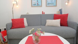 Un canapé gris avec des coussins rouges et blancs, un vase rempli de fleurs posé sur un set de table rouge, et deux tableaux encadrés accrochés au mur.