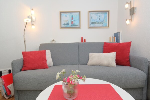 Een grijze bank met rode en witte kussens, een vaas met bloemen op een rode onderzetter, en twee ingelijste foto's aan de muur.
