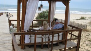 Een houten prieel met witte gordijnen, met uitzicht op een strand met een houten looppad.