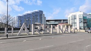 Wegwijzer met de tekst 'ARSENAL' in grote letters, een modern gebouw met glazen gevel en een helderblauwe lucht op de achtergrond.
