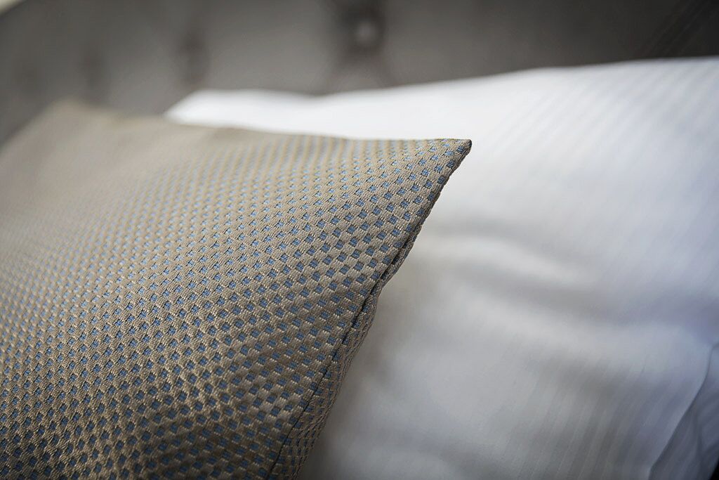 A close-up of a textured pillow with a geometric pattern, part of a bed with a white pillow and a grey headboard.