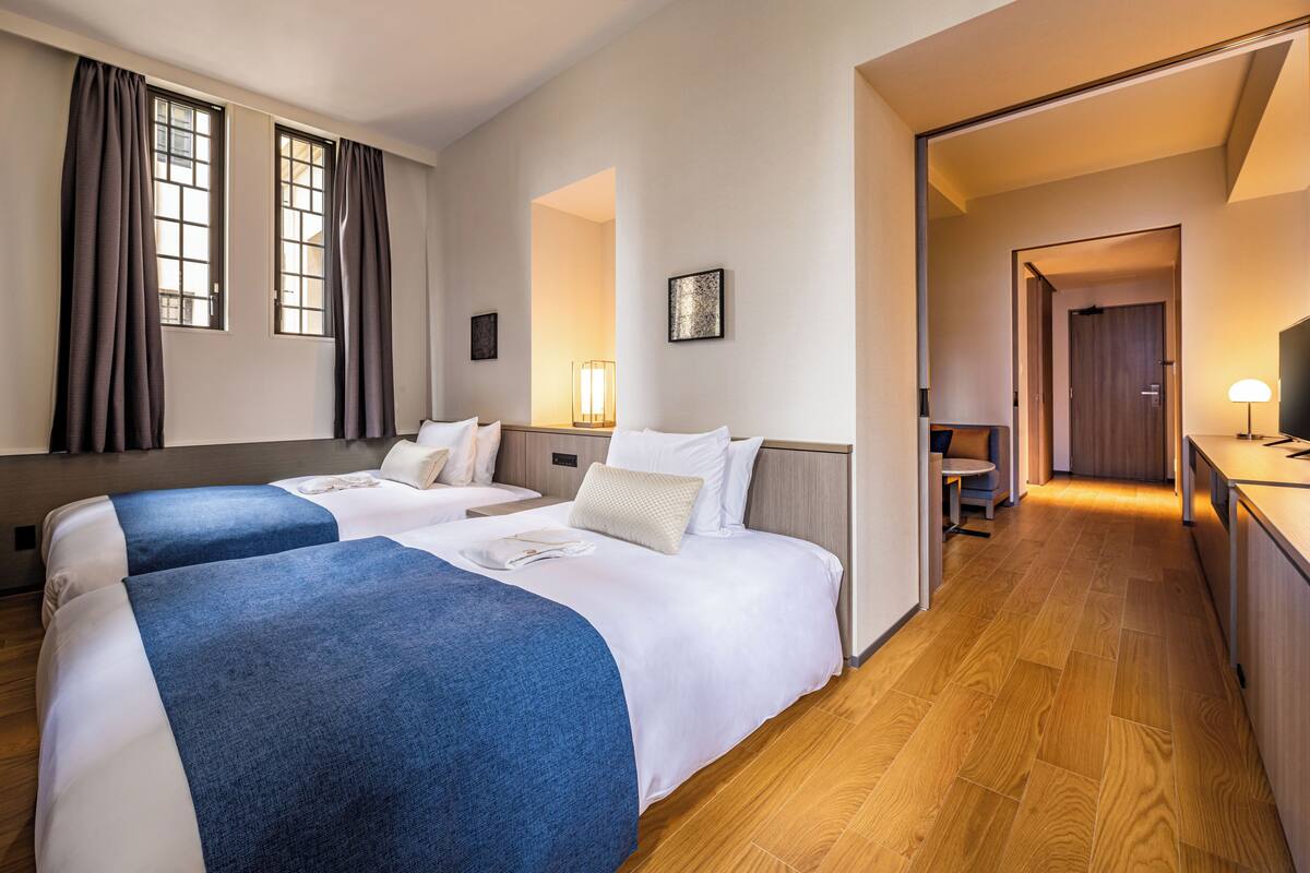 A hotel room with two beds, a desk, a chair, a lamp, and a door leading to another room.