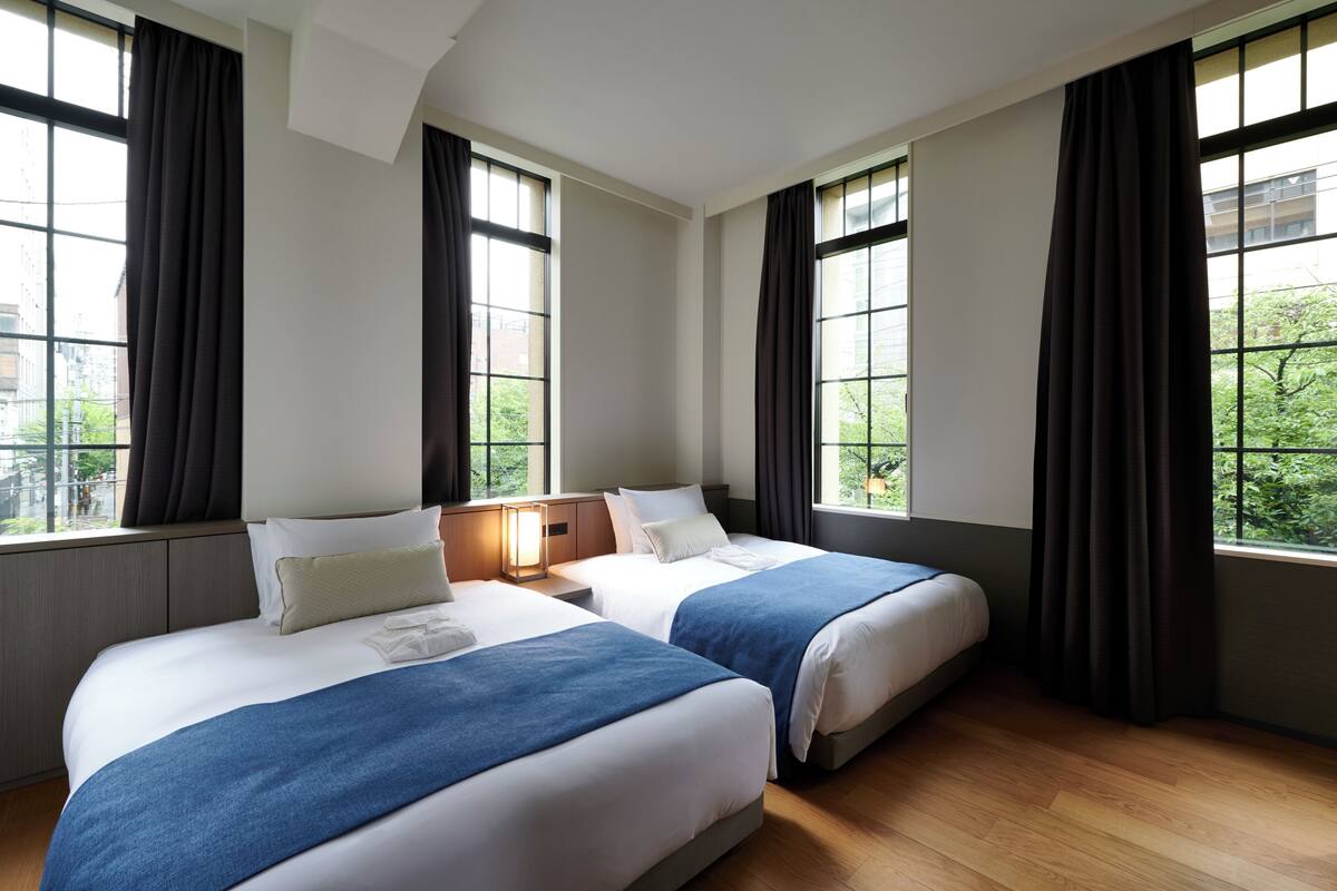 A hotel room with two beds, large windows, and a view of greenery outside.