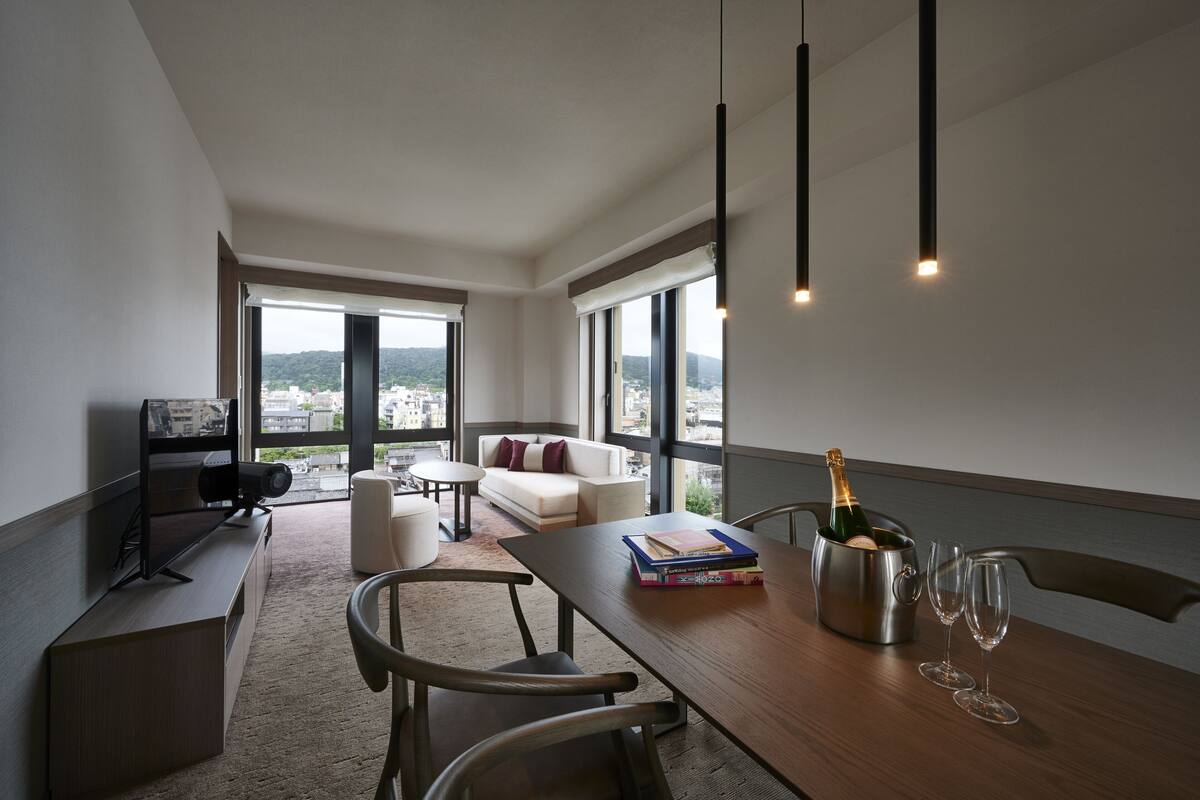 Una sala de estar moderna con una mesa de madera, libros, una botella de champán y copas de vino. Cuenta con un sofá, un televisor y ventanas amplias con vistas a la ciudad.