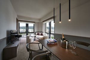A modern living room with a wooden table, books, a bottle of champagne, and wine glasses. There is a sofa, a TV, and large windows offering a city view.